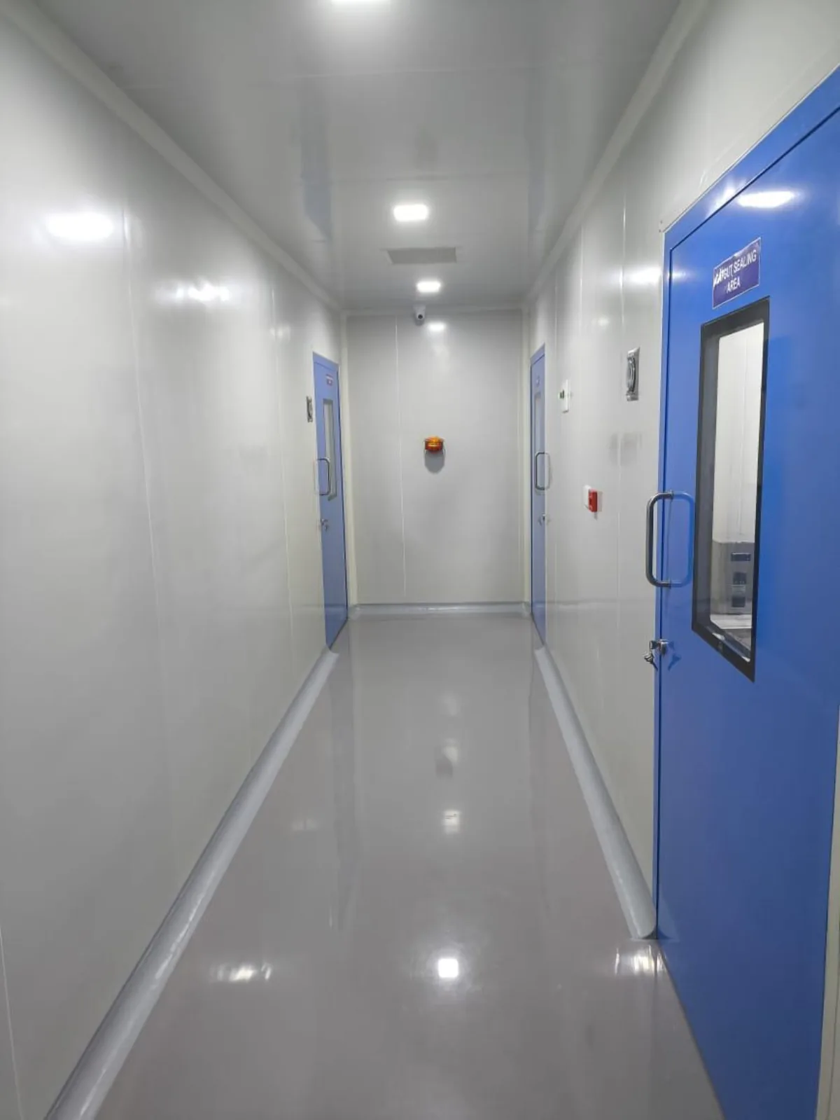 Cleanroom corridor with white modular wall panels, blue doors to the pouch sealing area, alarm beacon and pass box visible through the vision panel
